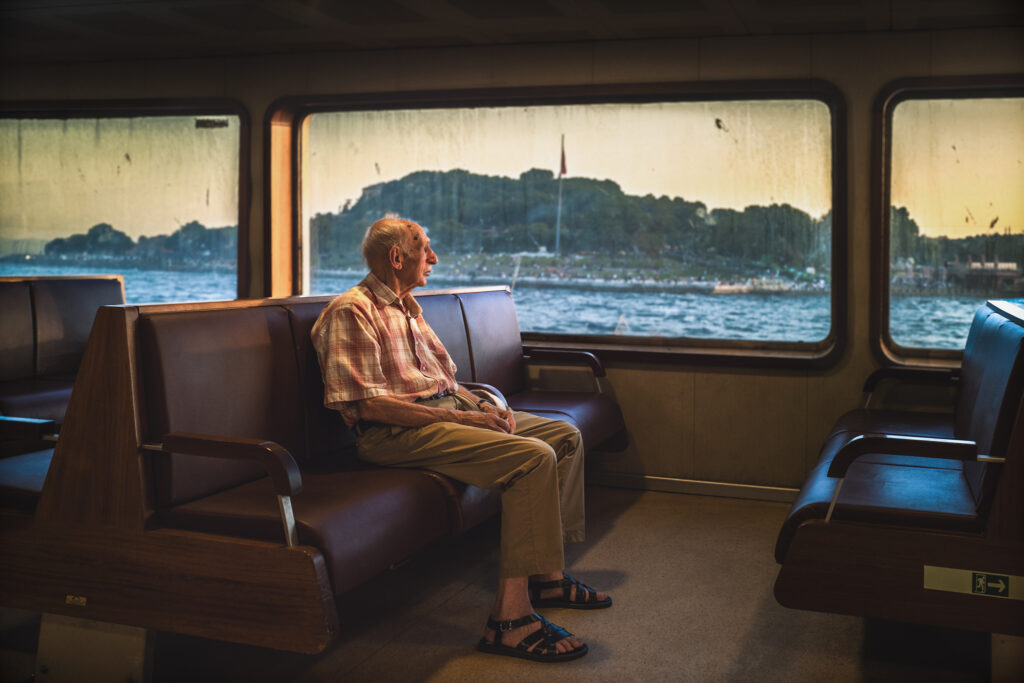 Imaginea surprinde un bătrânel privind cu melancolie pe fereastra feribotului, cu fața luminată de razele soarelui care pătrund prin sticlă. Îmbrăcămintea sa simplă și poziția mâinilor împreunate îi oferă un aer de nostalgie, iar privirea lui pare să caute ceva în depărtare. Pe fundal se văd valurile mării și siluete ale peisajelor îndepărtate, care se conturează la orizont. Totul în jurul său sugerează o liniște profundă, în timp ce feribotul se îndreaptă spre un nou orizont.