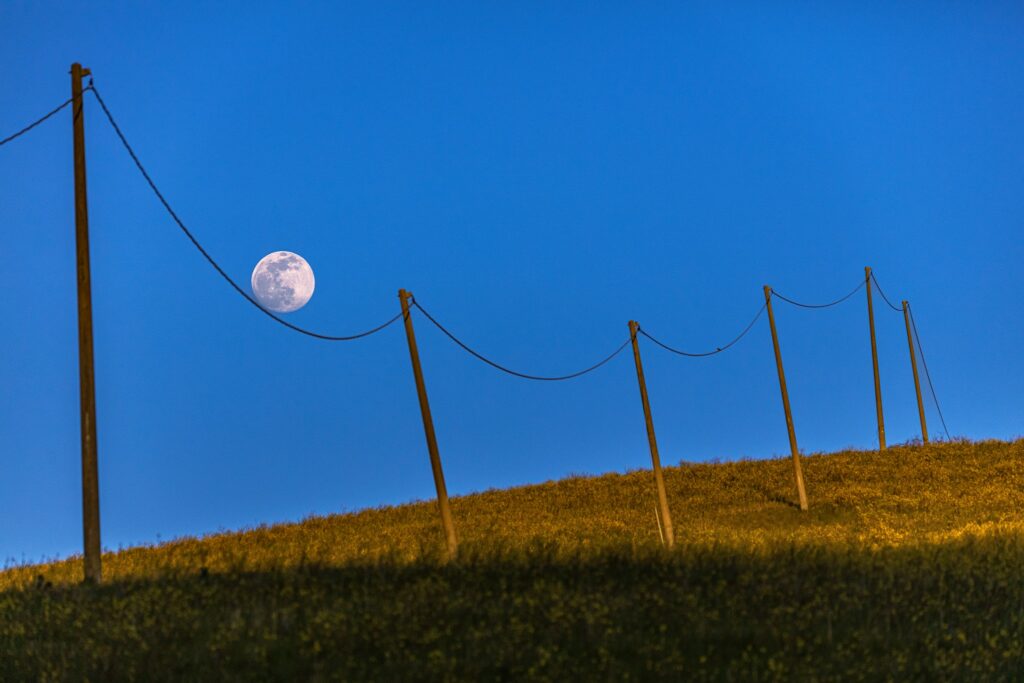 Cer perfect senin, albastru deschis intens pe care strălucește o lună perfect circulară, deși încă e multă lumină afară. Cam un sfert din poză se află și un deal pe care sunt niște stâlpi înalți de electricitate, cu cabluri precum niște acolade. Unul dintre aceste cabluri încadrează partea de jos a lunii, făcând-o să pară că alunecă în jos pe sfoară.