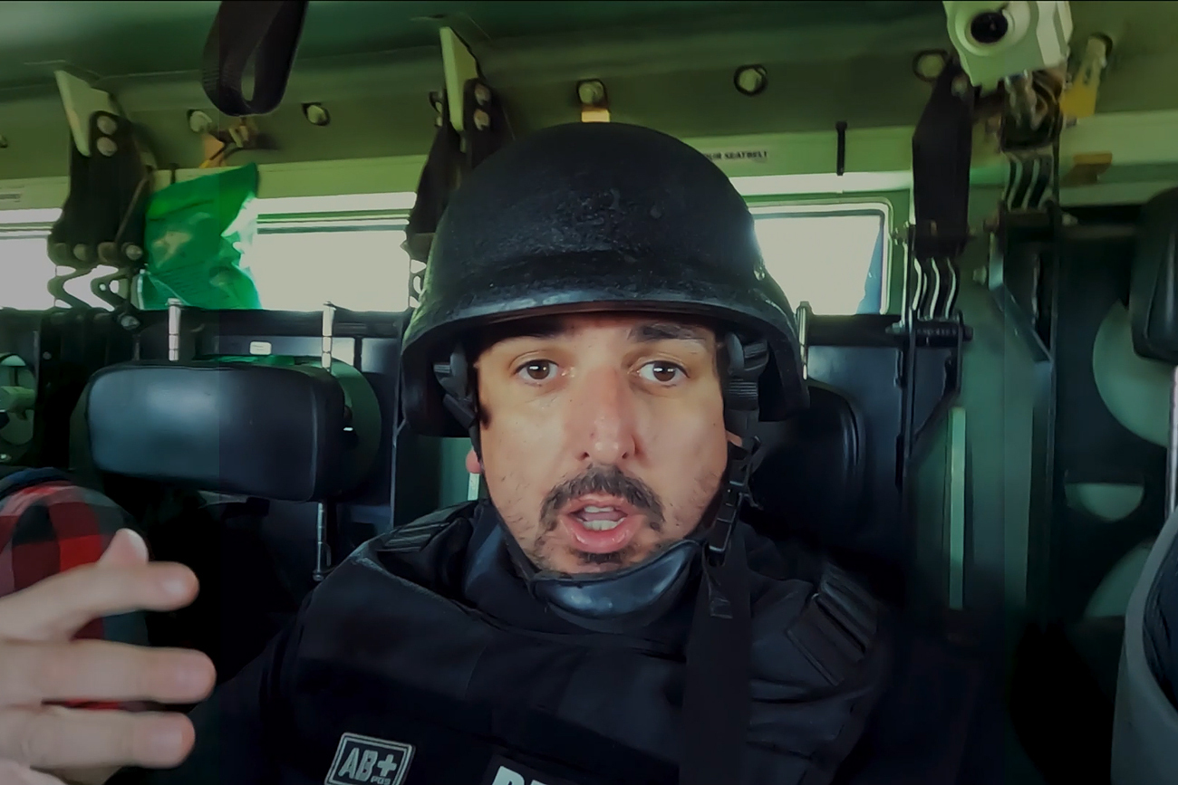 Portret cu Mircea Barbu, jurnalist în zone de conflict, în interiorul unei dube, cu echipament de protecție (cască și vestă), vorbind și gesticulând spre cameră.