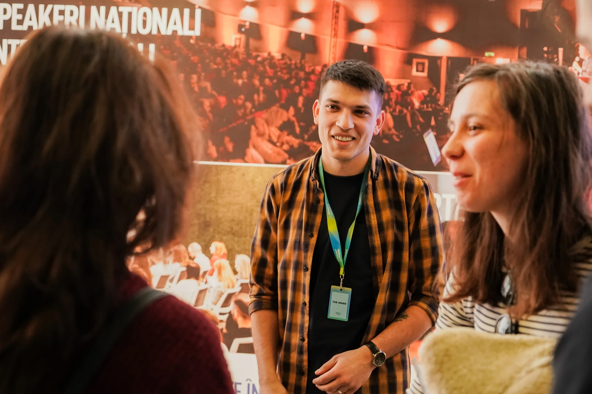 Vlad Guluță menorând tineri la un eveniment, în cămasă cu carouri, cu zâmbet pe față și uitându-se cu empatie spre doi tineri care discută.