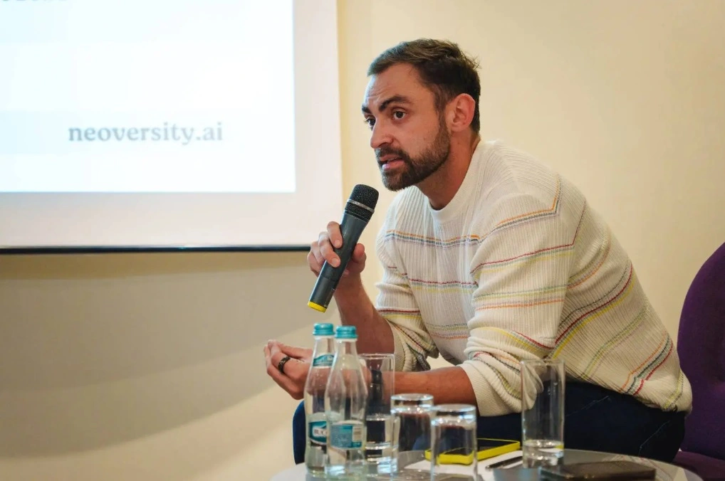 Portret cu Tinu, având în spate un ecran cu o prezentare, microfon în mână și vorbind stând pe scaun.