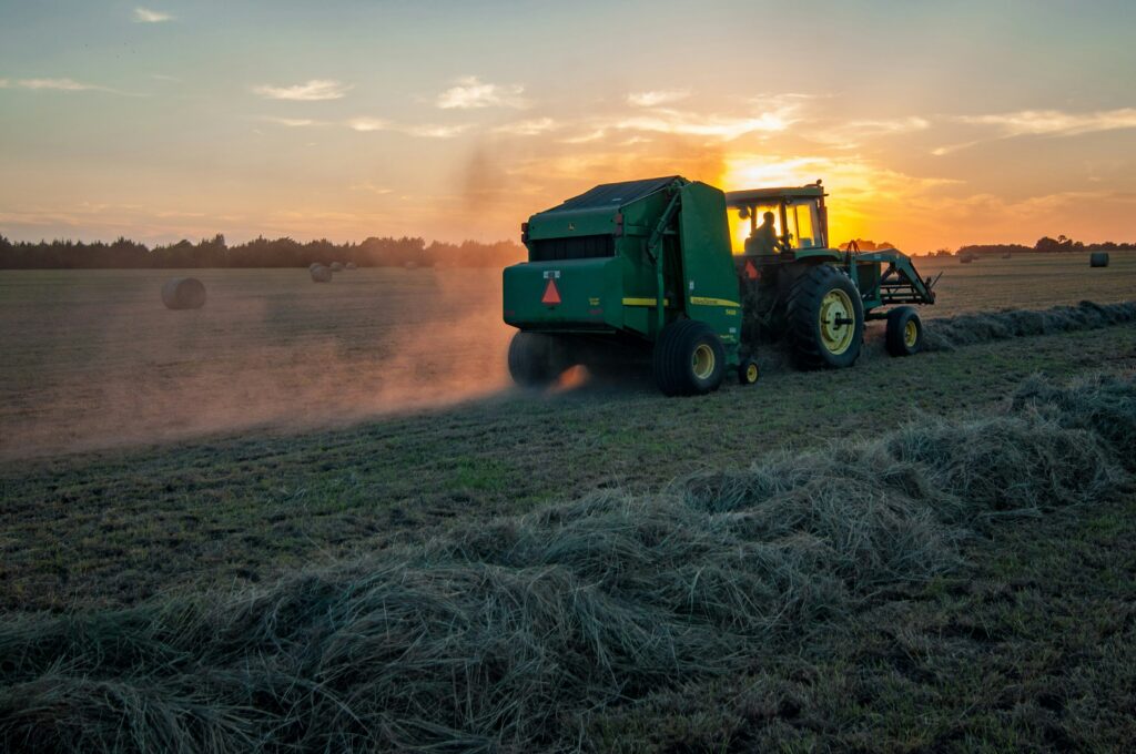 Un fermier cu un tractor verde pe un câmp de iarbă cosită, la apus.