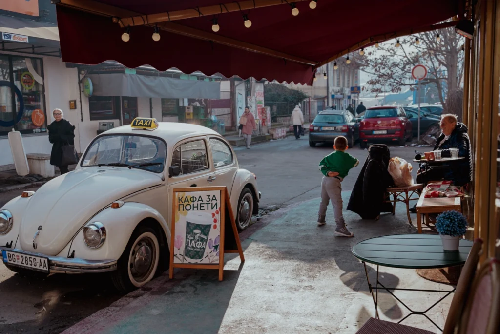 Fotografie de stradă, un taxi vechim un copil vesel și doi bătrânei în fața unei cafenele.