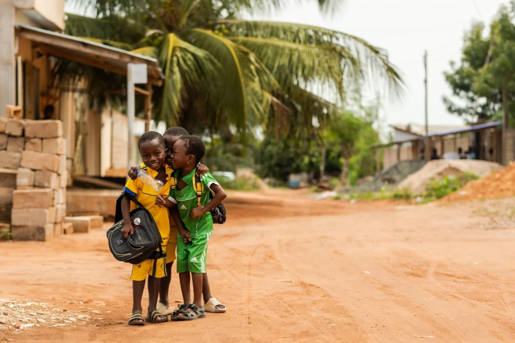 Trei copii africani îmbrăcați colorat cu verde și galben care se uită curioși spre fotograf și șușetesc entuziasmați.