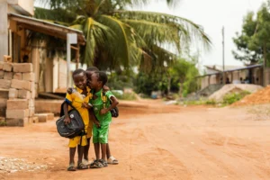 Trei copii africani îmbrăcați colorat cu verde și galben care se uită curioși spre fotograf și șușetesc entuziasmați.