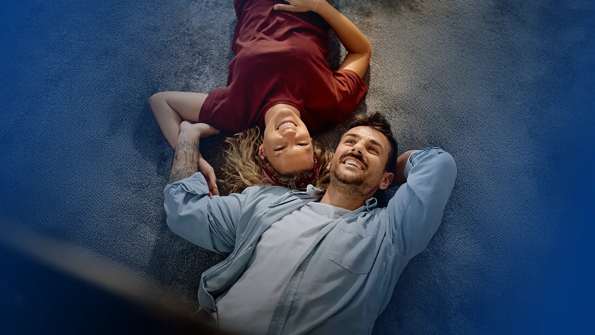 Un cuplu tânăr stă relaxat pe podea, zâmbind și privind în sus, sugerând un moment de liniște, siguranță și visare pentru viitor