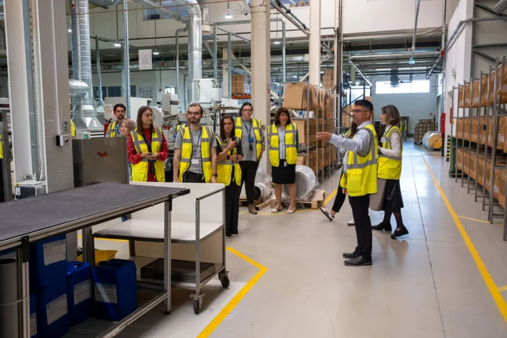 Un grup de angajați poartă veste reflectorizante galbene în timpul unui tur ghidat în hala de producție Rottaprint, printre rafturi și echipamente industriale